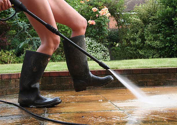 Jeune femme en bottes en caoutchouc en train de nettoyer sa terrasse en carrelage avec un jet d'eau